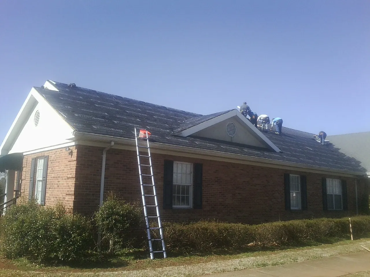 Crew tearing shingles off a brick ranch roof with a ladder set — Emergency Roof Repair work by our crew in Dola