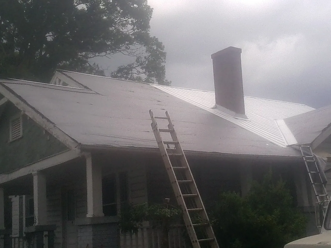 Ladders against a metal roof on an older home with a brick chimney in Dola