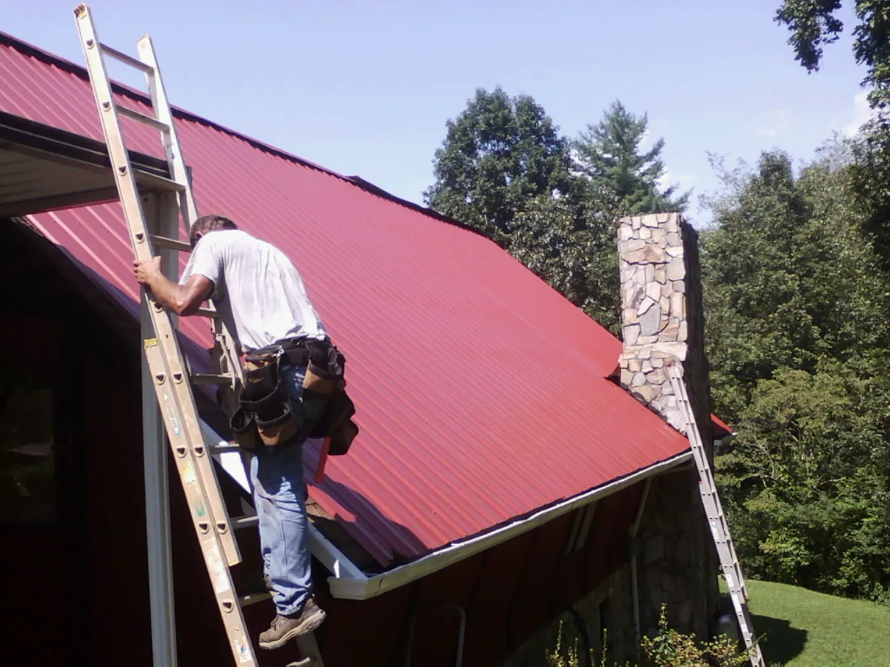 Roofer on a ladder beside a red metal roof and stone chimney — Soffit & Fascia Repair by Roofing Brothers in Dola